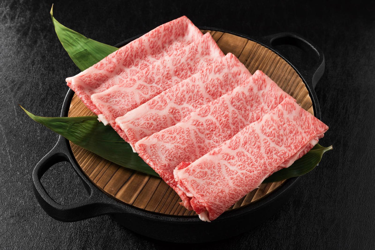 Folded marbled wagyu slices on a wooden board in a black dish with green leaves