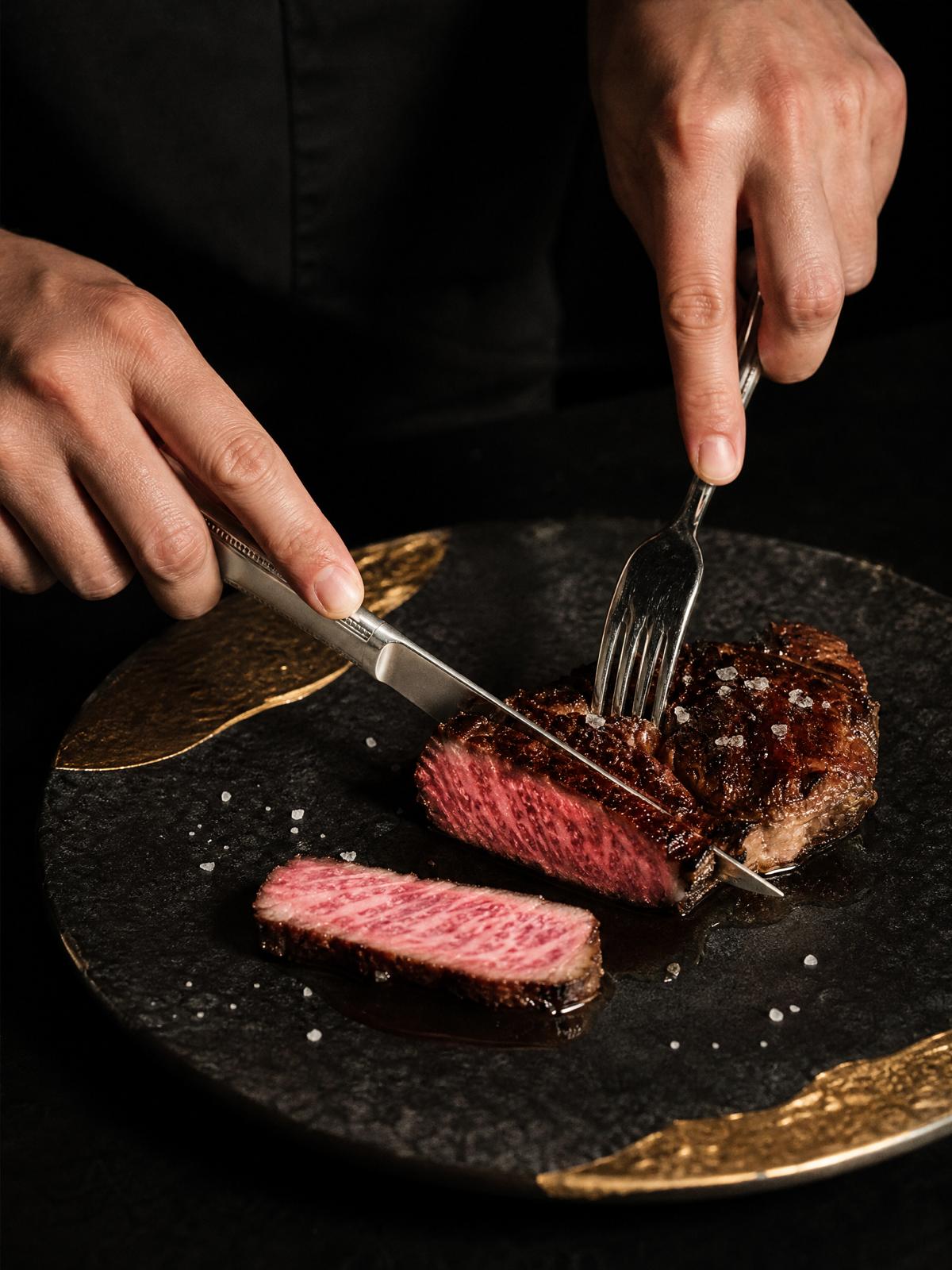 Hands slicing a seared steak with knife and fork on a dark, gold-edged plate